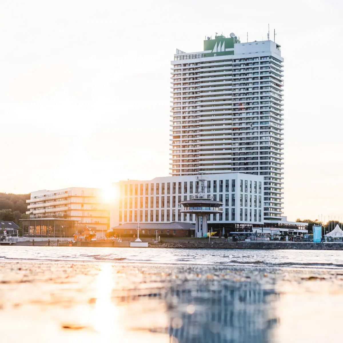 Ein großer quadratischer Hotelkomplex mit vielen Fenstern direkt am Meer. Die Morgensonne strahlt voller Kraft hinter dem Hotel hervor und spiegelt sich im flachen Wasser.