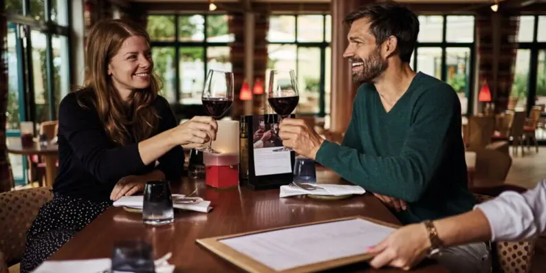 Eine Frau und ein Mann sitzen in einem Bistro und stoßen mit einem Glas Rotwein an.