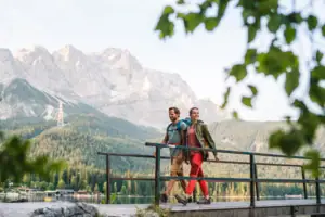 Wandern Ein Paar spaziert auf einer Brücke mit einem See und Bergen im Hintergrund.