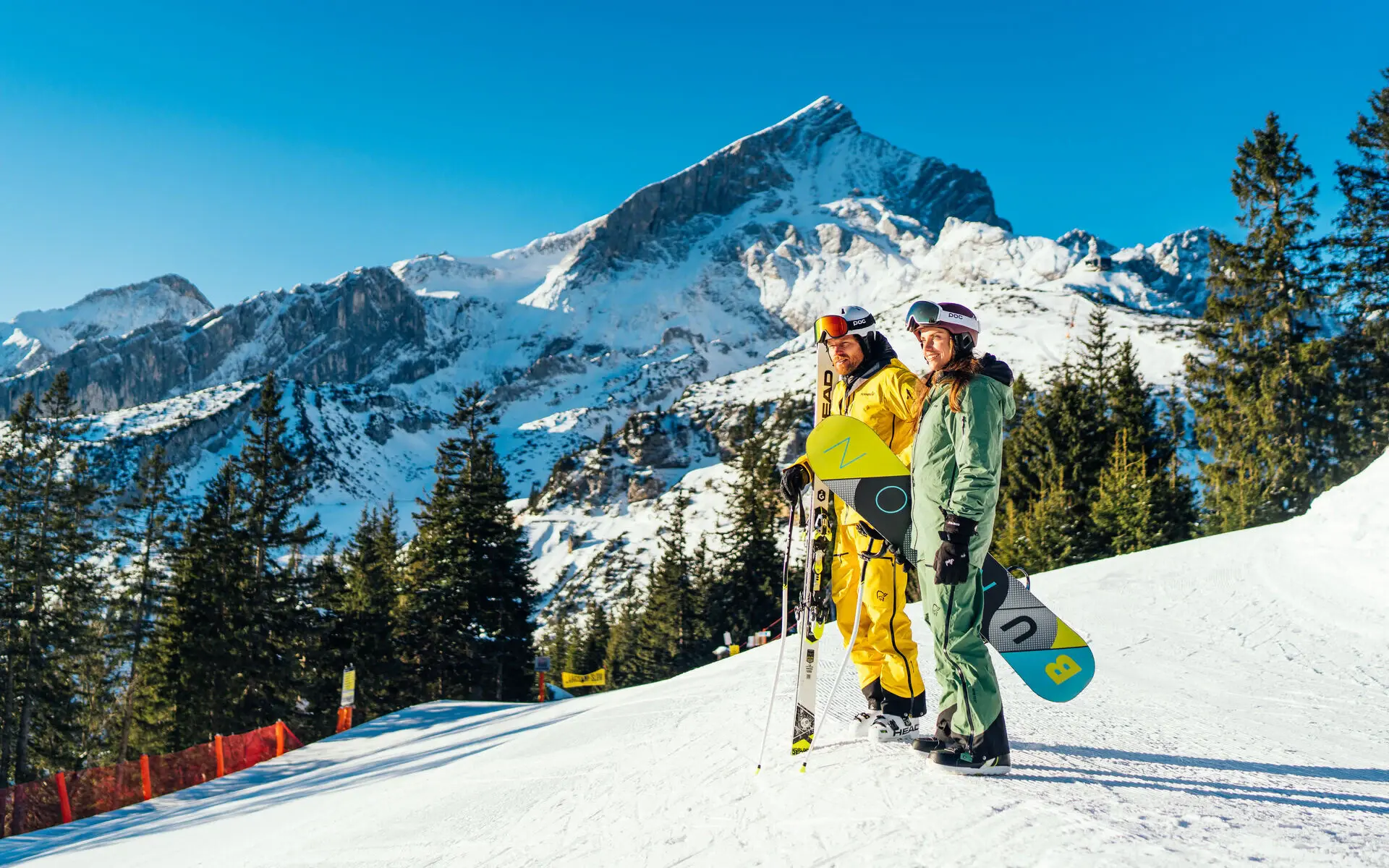 Zwei Personen stehen mit Skiausrüstung auf einem Berg mit Schnee, im Hintergrund ist eine schneebedeckte Berglandschaft