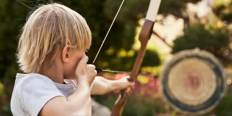 A blond boy draws a bow and aims at a target.