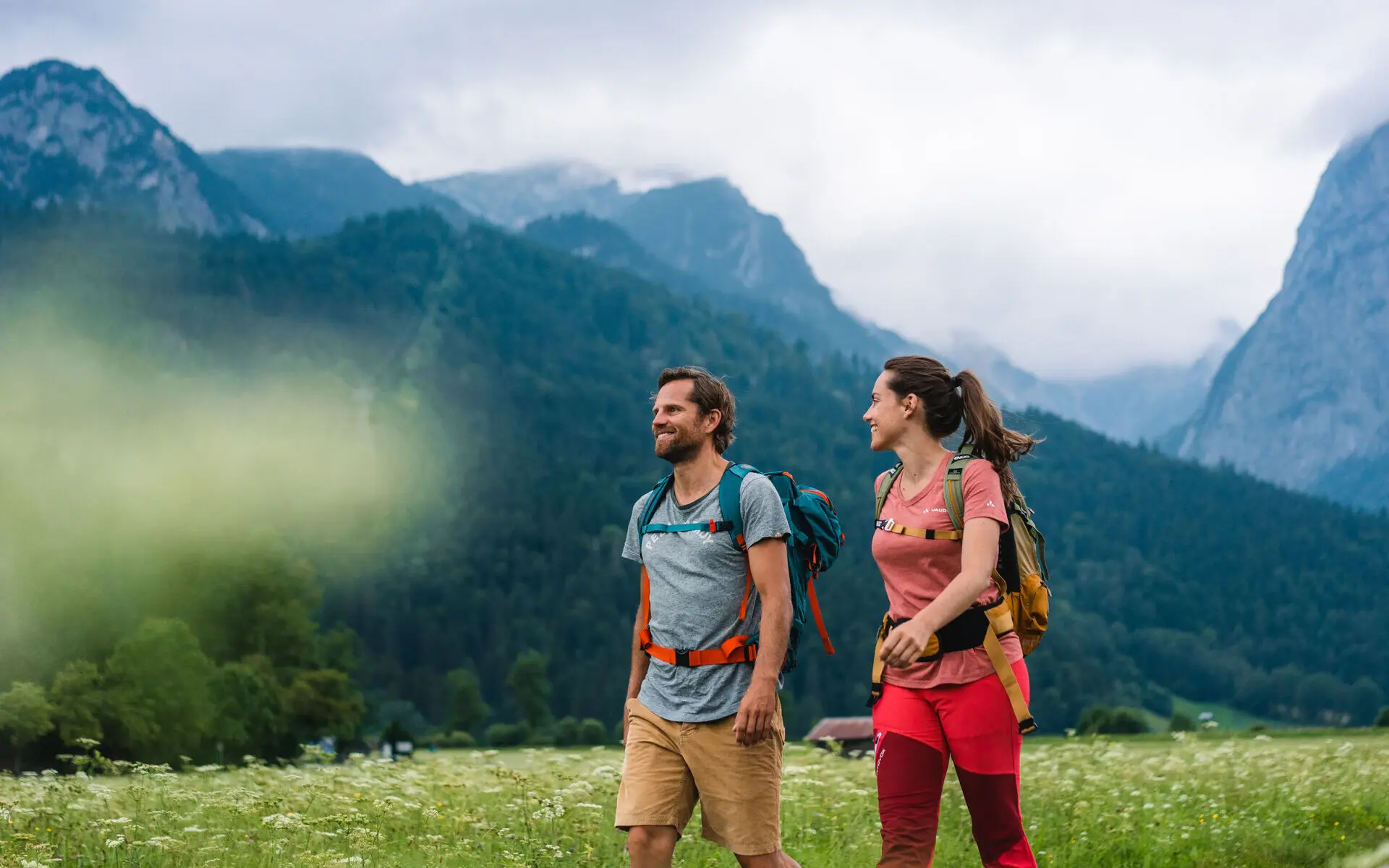 Ein Mann und eine Frau spazieren auf einem Pfad durch ein Feld, im Hintergrund sind Berge zu sehen.