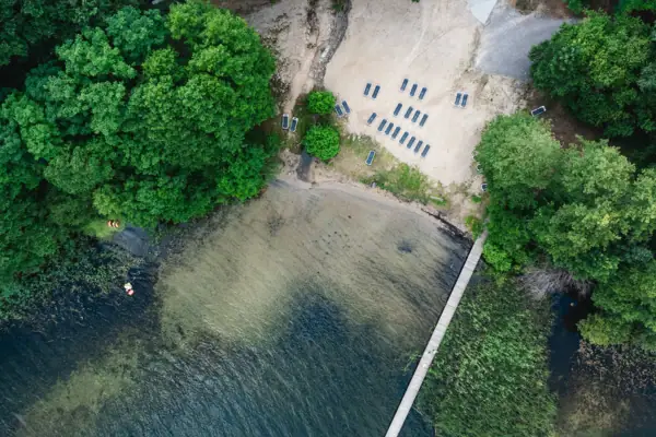 Scharmützelsee Badestelle Ein Steg führt über einem See neben einem Strand mit Stühlen und Bäumen.