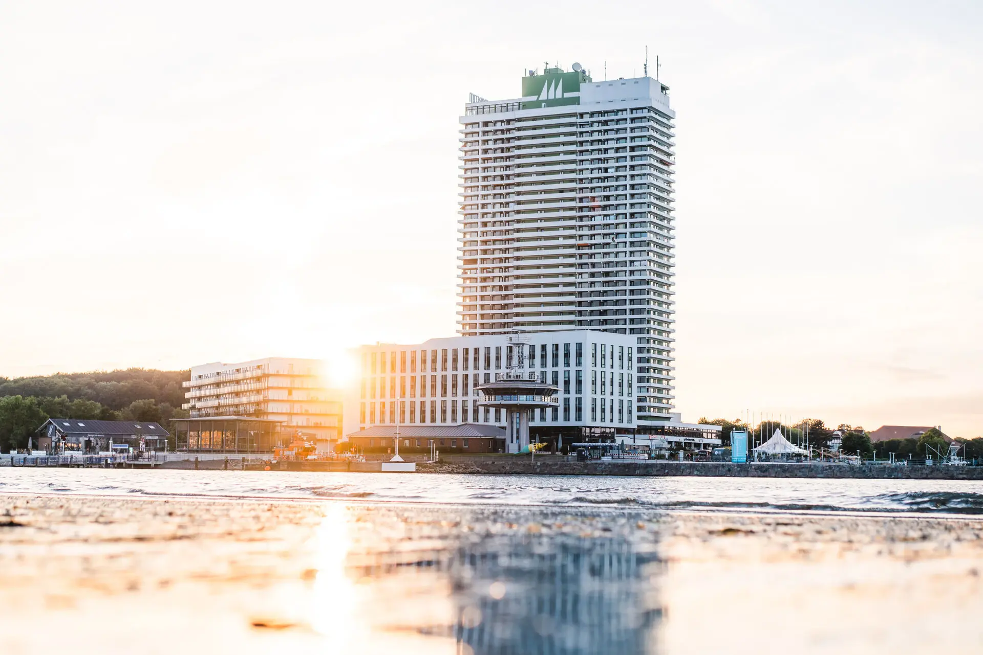 Ein großer quadratischer Hotelkomplex mit vielen Fenstern direkt am Meer. Die Morgensonne strahlt voller Kraft hinter dem Hotel hervor und spiegelt sich im flachen Wasser. 