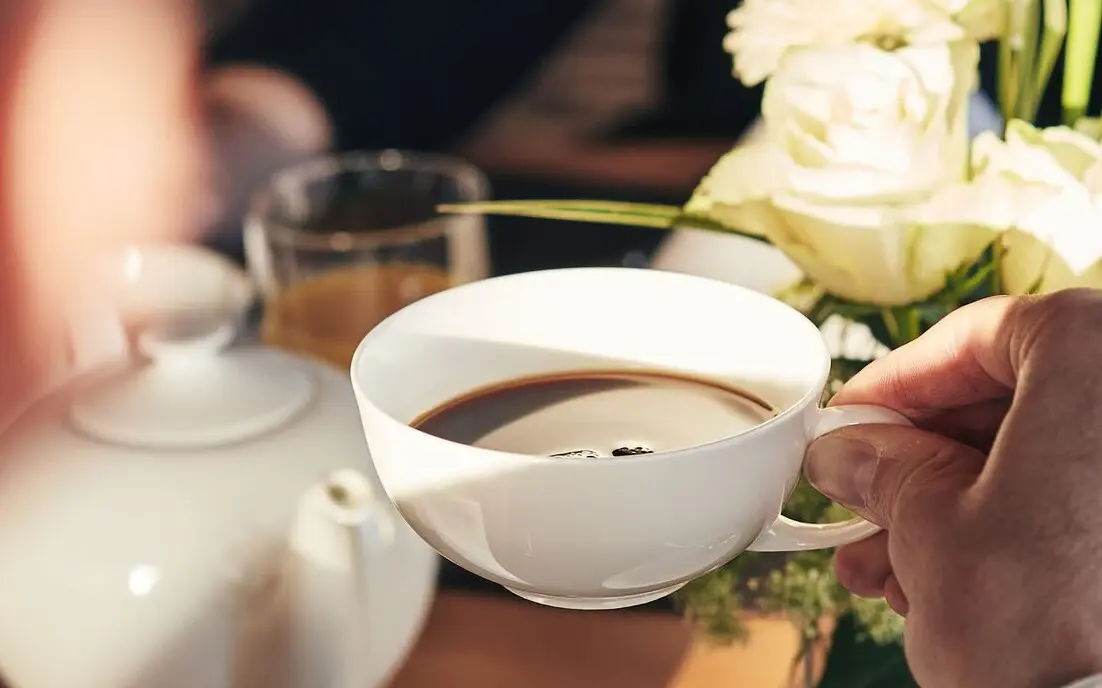 Person hält eine Tasse Kaffee und im Hintergrund ist eine Kanne und Rosen zu sehen.