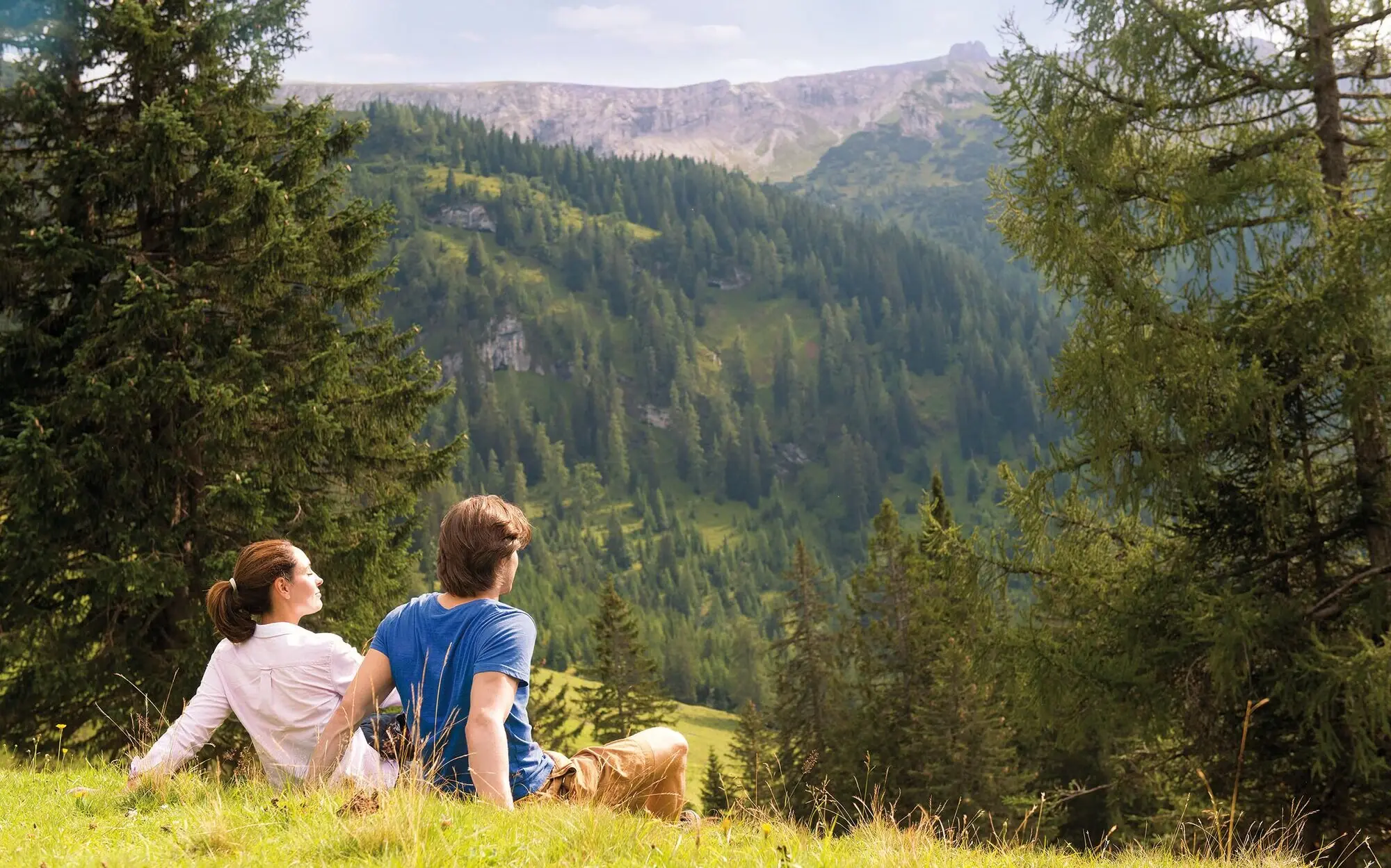 Ein Mann und eine Frau sitzen auf einem Hügel und blicken auf ein Bergpanorama.