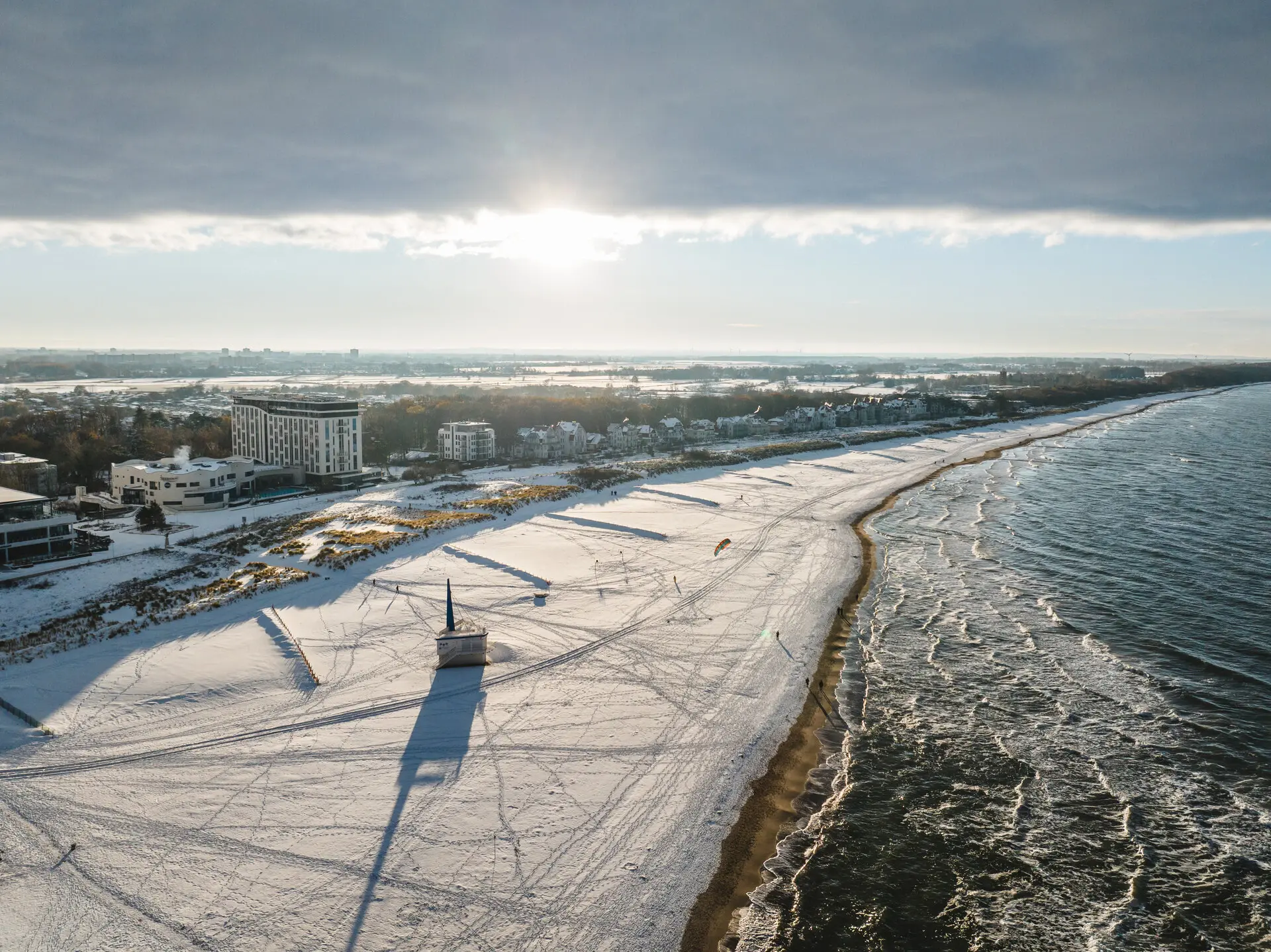 Winterliche Szenerie eines schneebedeckten Strandes mit Gebäuden und Wasser.