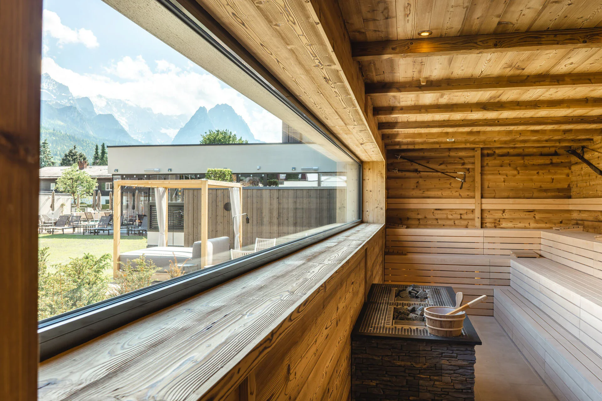 Hölzerner Saunaraum mit Saunaofen vor einem Fenster in einer Holzwand mit Blick auf die Berge