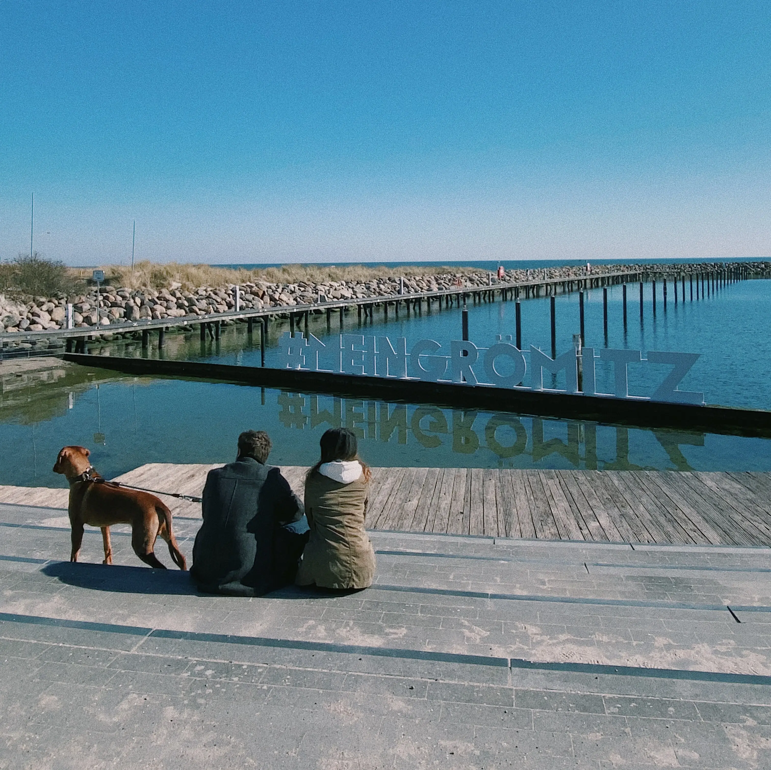 Menschen sitzen mit einem Hund auf einem Dock und schauen aufs Wasser.