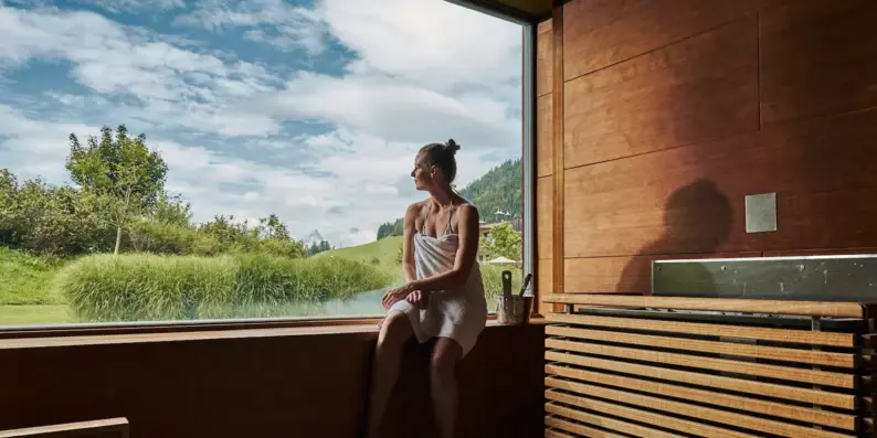 Frau sitzt in einer Sauna auf einer Fensterbank und blickt durch ein großes Panoramafenster nach draußen.