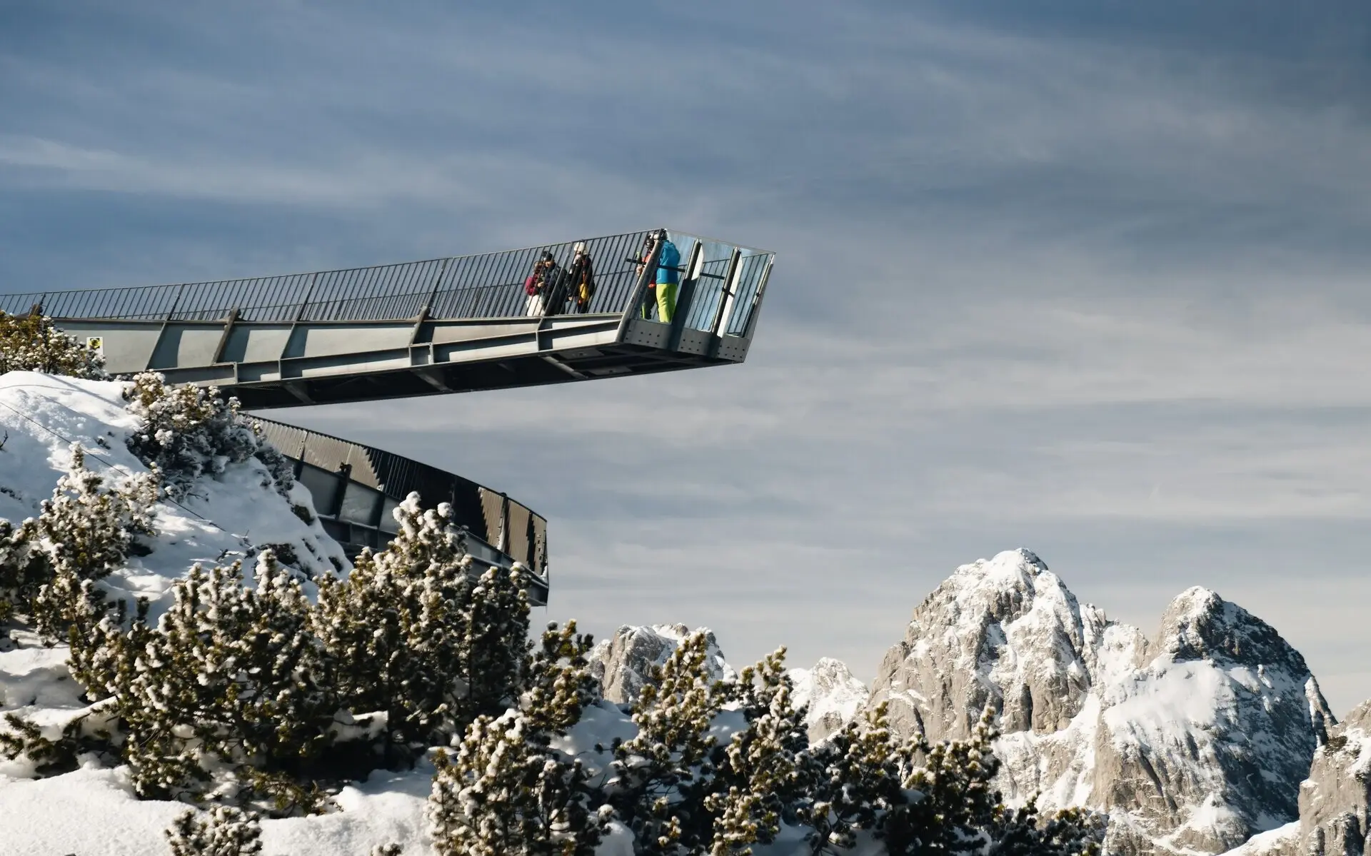 Personen gehen über eine Aussichtsplattform in einer verschneiten Berglandschaft.