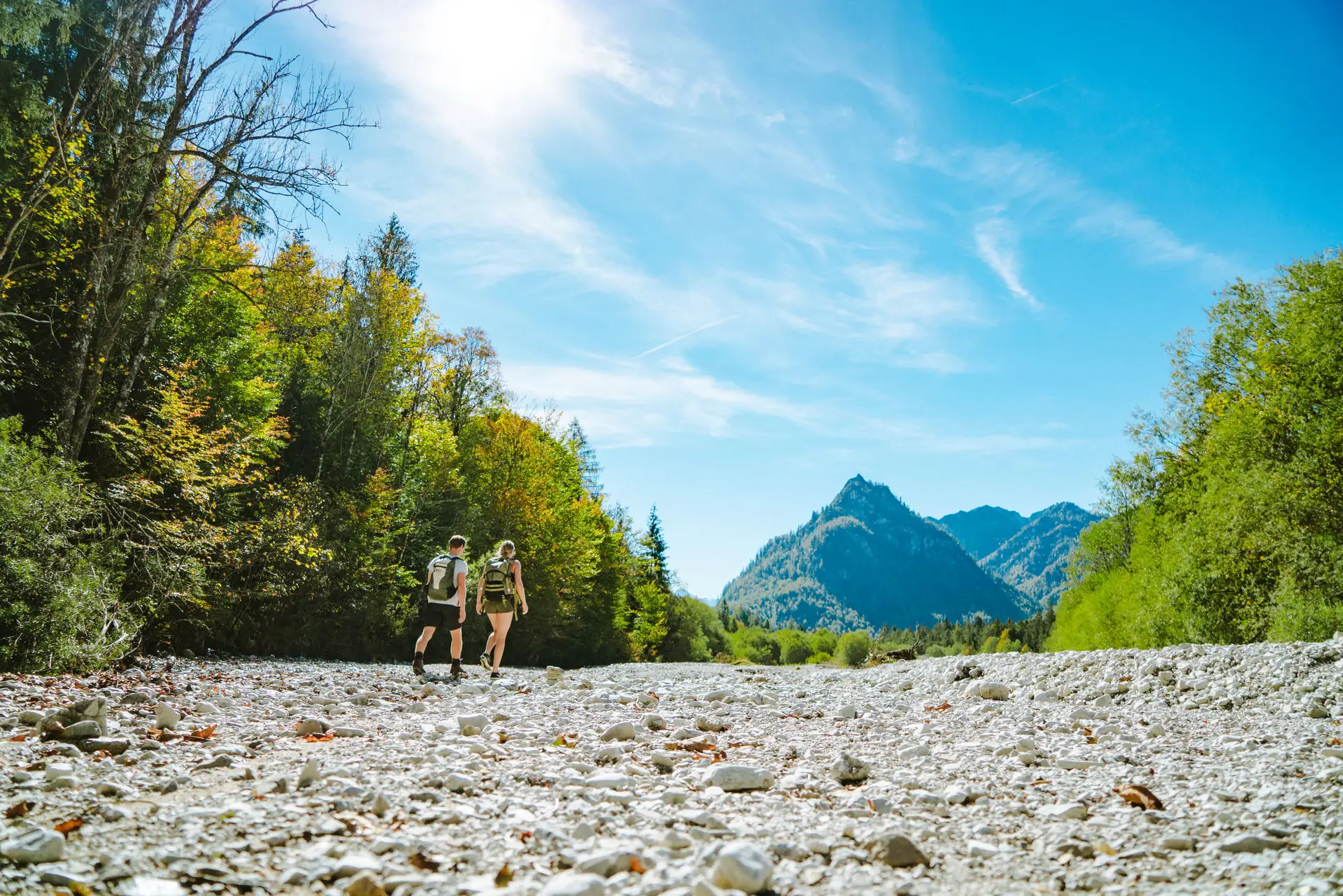 Wanden durchs Flussbett Eine Gruppe von Menschen wandert auf einem felsigen Pfad mit Bäumen und Bergen im Hintergrund.