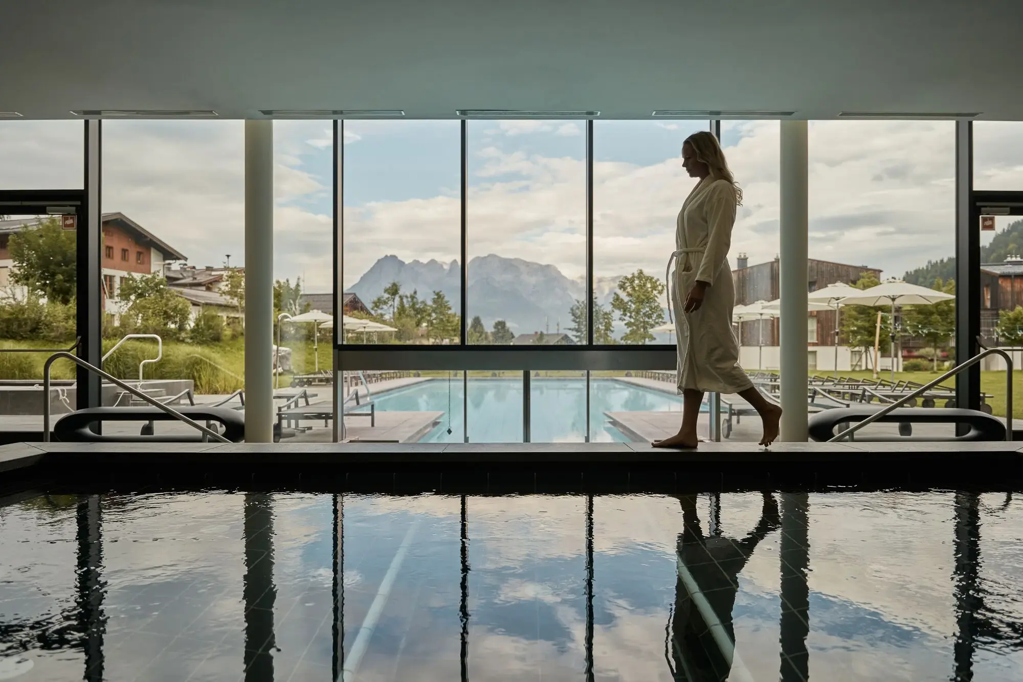 Frau geht an einem  Innenpool vorbei und im Hintergrund ist der Außenpool und eine schöne Bergkulisse zu sehen.