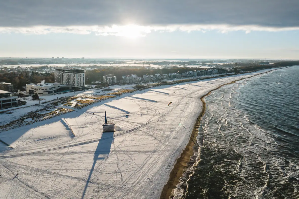 Winter Warnemünde Schneebedeckter Strand mit Gebäuden und Wasser im Winter.