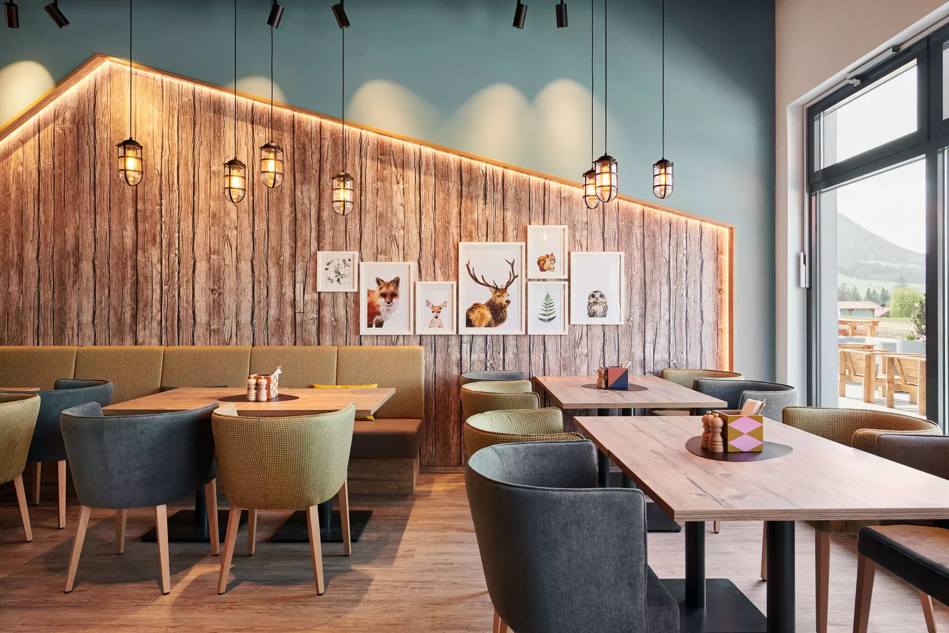 Stilvoll eingerichteter Restaurantbereich mit Holzwand und gemütlichen Sitzgelegenheiten. An der Wand hängen Bilder mit Tiermotiven, die dem Raum eine warme, naturverbundene Atmosphäre verleihen. Pendelleuchten und eine dezente Hintergrundbeleuchtung sorgen für eine einladende, entspannte Stimmung.