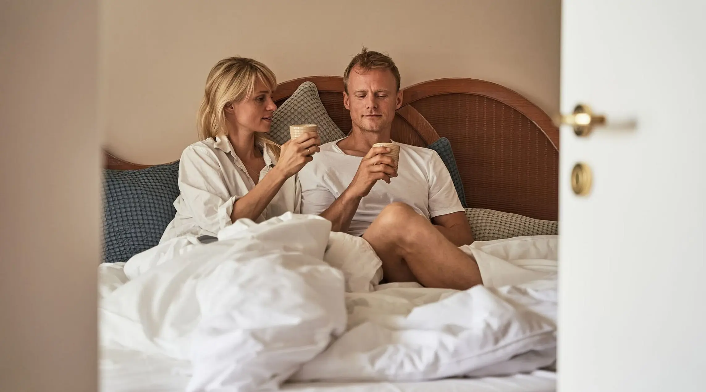 A man and a woman sit on a bed and hold cups in their hands.