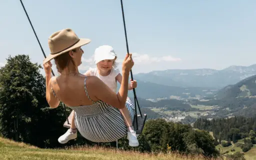 Eine Mutter und ihr Kind schaukeln gemeinsam auf einer Wiese mit Blick auf die idyllische Landschaft von Ruhpolding, umgeben von Bergen und strahlend blauem Himmel. 