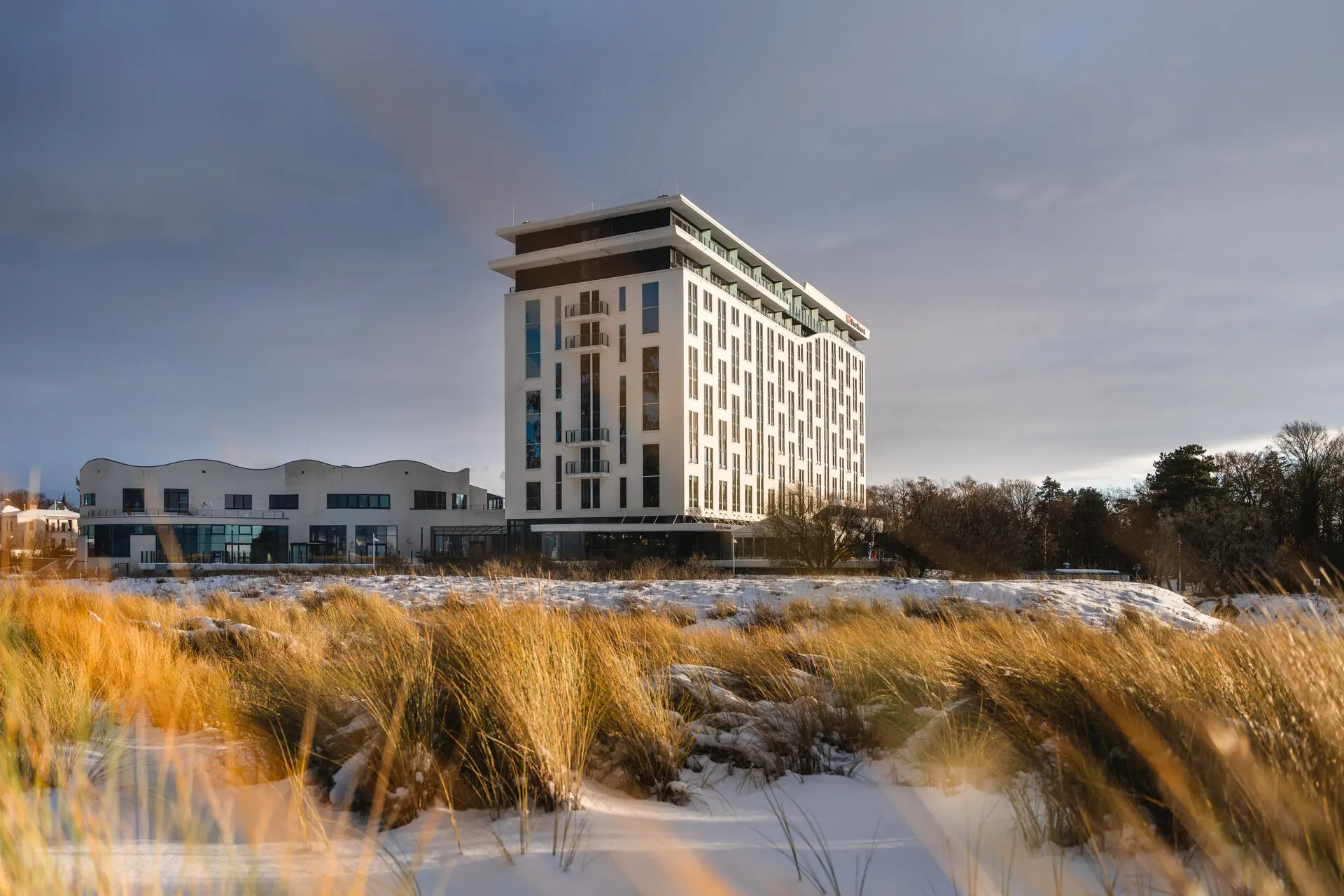 Ein modernes Hotelgebäude in Küstenlage, umgeben von winterlicher Landschaft mit schneebedecktem Sand und hohen Gräsern, unter einem bewölkten Himmel