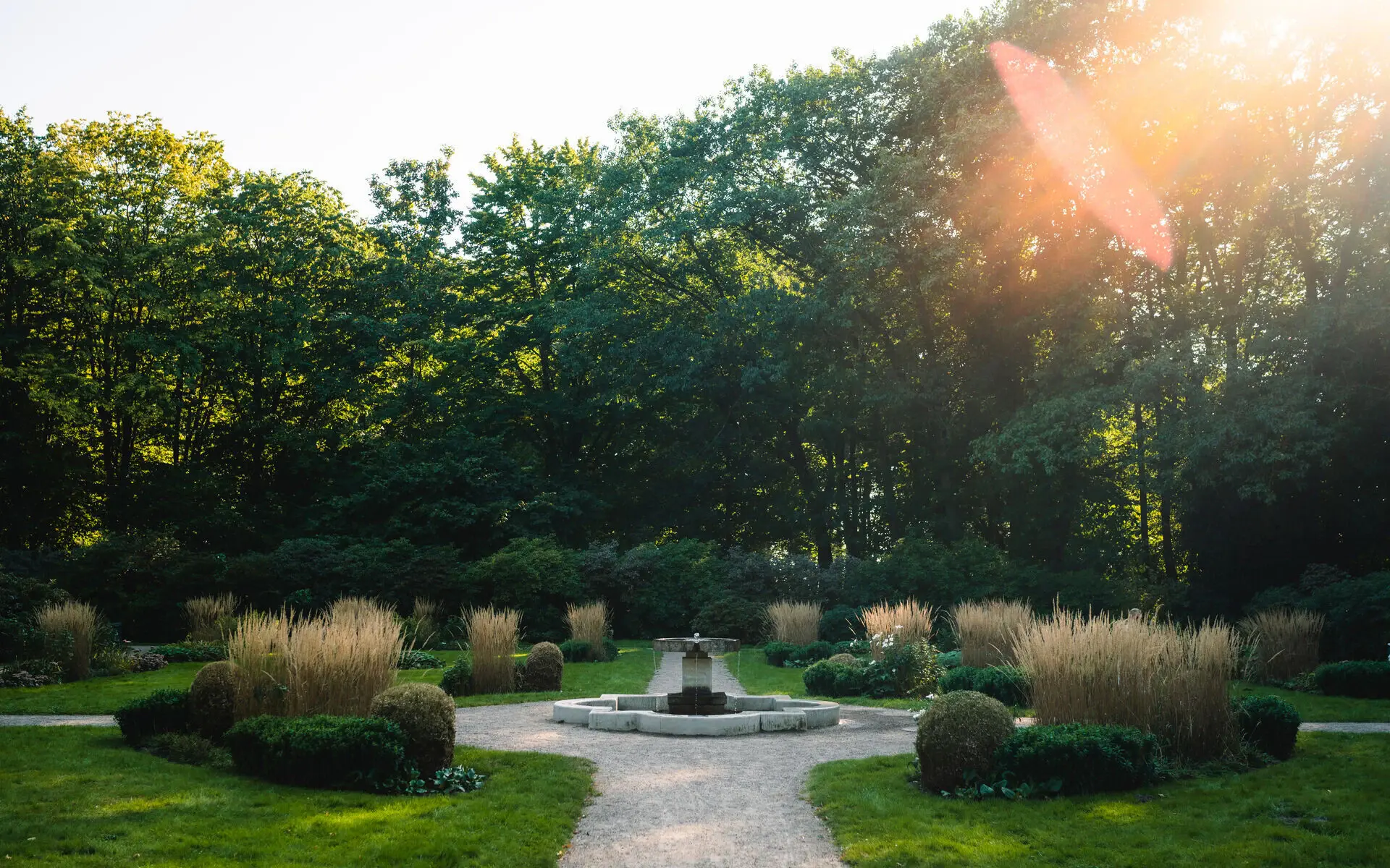 Ein Park mit kleinem Springbrunnen bei aufgehender Sonne, welche durch die umstehenden Bäume fällt. 