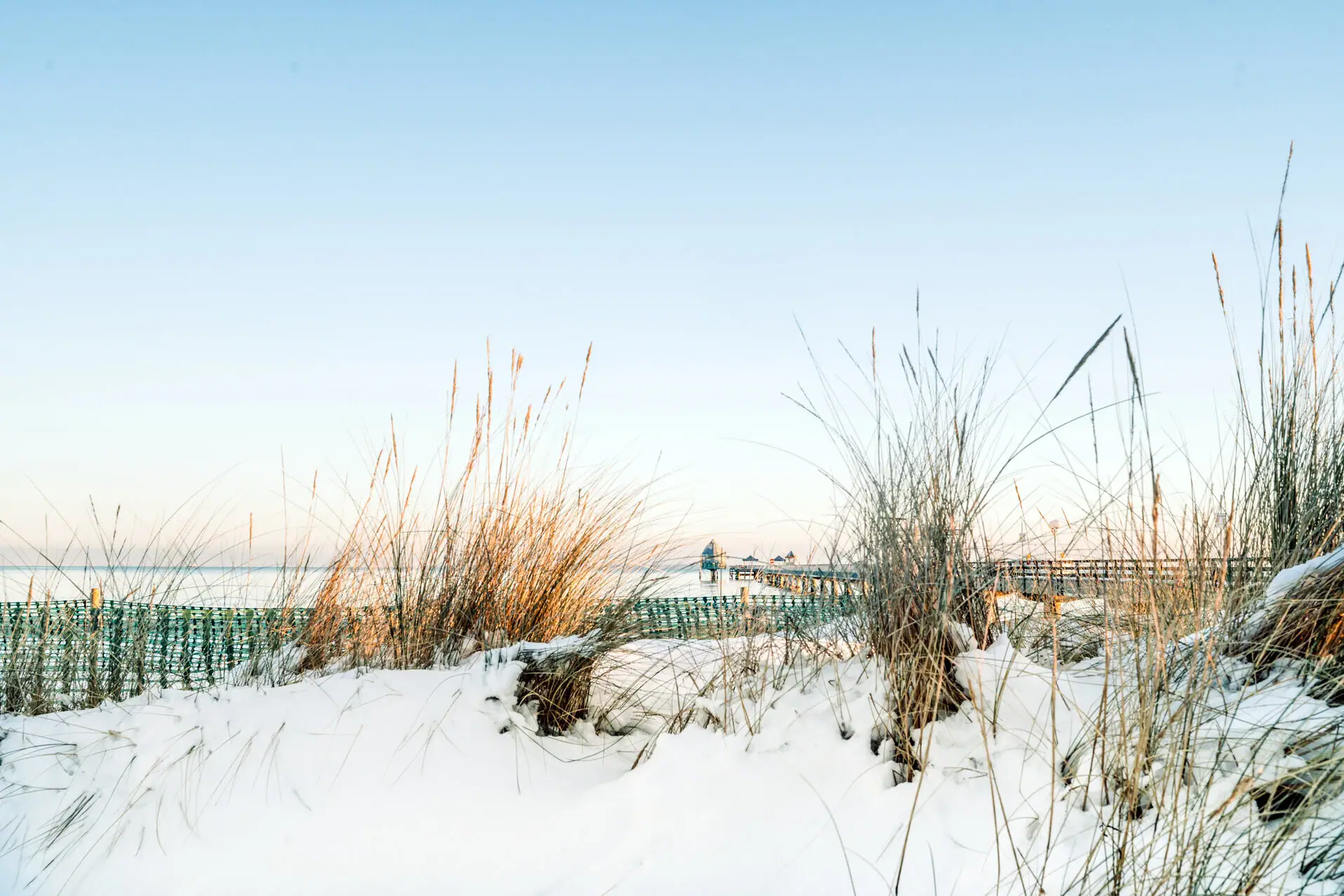 Schneebedeckter Boden mit hohem Gras und einem Pier im Hintergrund.