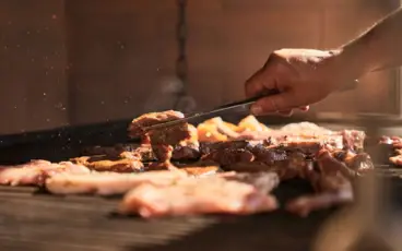 Eine Nahaufnahme von Fleisch, welches auf einem Grill gebraten wird. Zudem ist eine Hand zu sehen, die eine Zange hält und ein Stück Fleisch wendet.