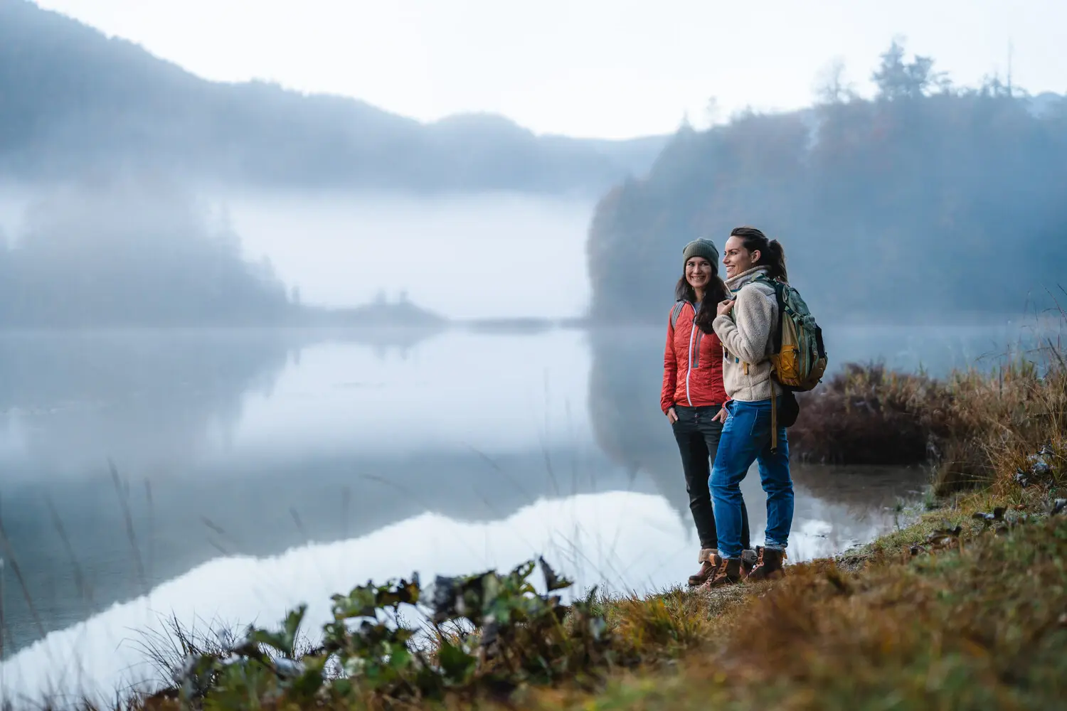 Zwei Frauen stehen neben einem See, umgeben von Nebel und Bergen.
