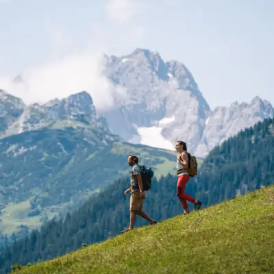 Zwei Personen wandern auf einem Hügel mit Bergen im Hintergrund.