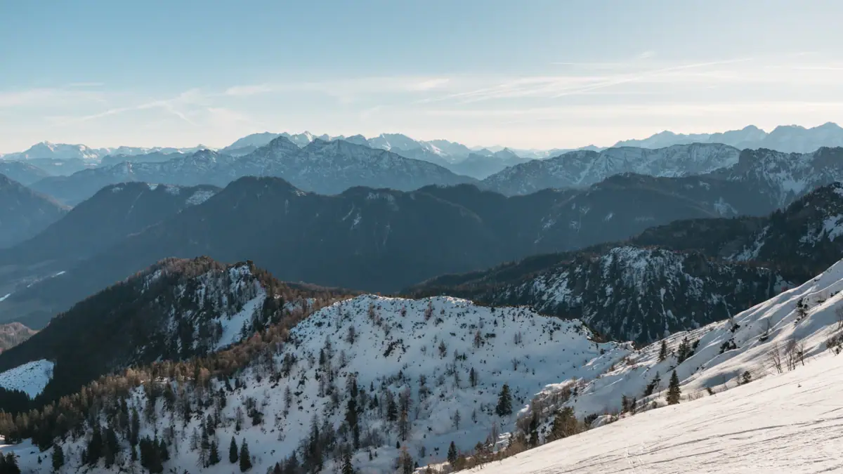 Verschneiter Berg mit Bäumen und weiteren Bergen im Hintergrund.