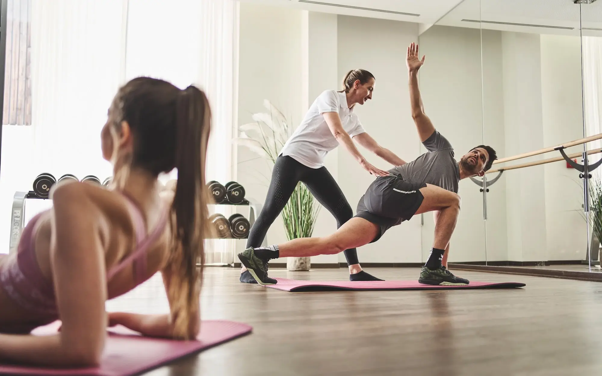 A man stretches on a yoga mat in a classroom while a woman gives him instructions and assistance.