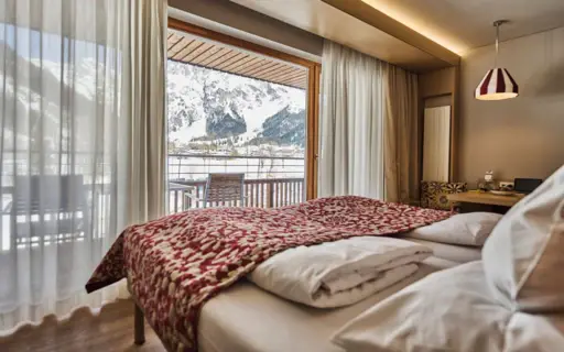 Ein Bett mit roter Decke in einem Zimmer, im Hintergrund ein Fenster mit Blick auf verschneite Berge.