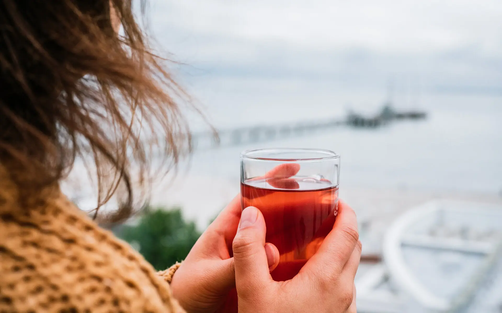 Eine Frau in senffarbenem Strickpullover steht bei windigem Wetter auf einer Terrasse und hält ein Glas Tee in ihrer Hand, während sie auf die Weiten des Meeres schaut. 