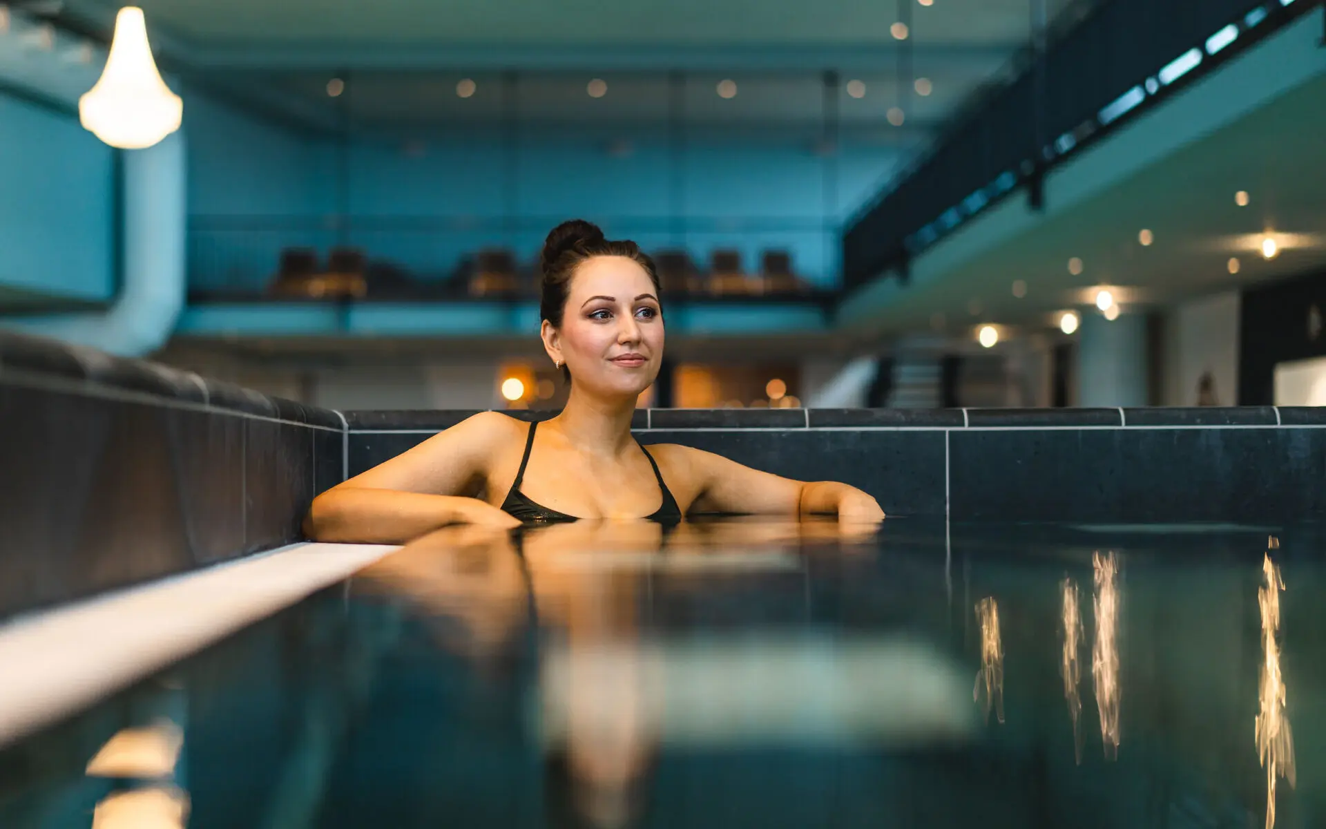 Eine dunkelhaarige Frau entspannt in einem eckigen Schwimmbecken, die Ellenbogen auf dem Beckenrand aufgestützt. Im Hintergrund ist abgedunkelt und leicht verschwommen der Schwimmbereich zu erkennen.