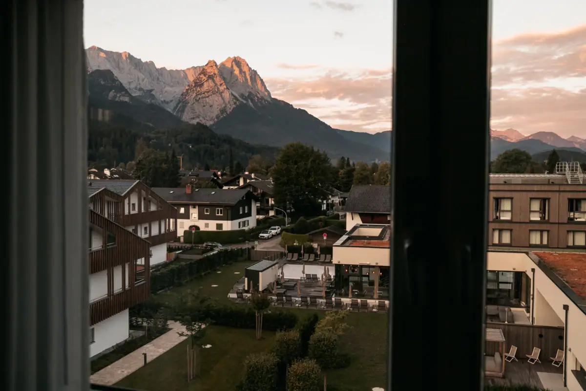 Bergblick Blick auf eine Stadt durch ein Fenster mit Bäumen, Wolken und Bergen im Hintergrund.