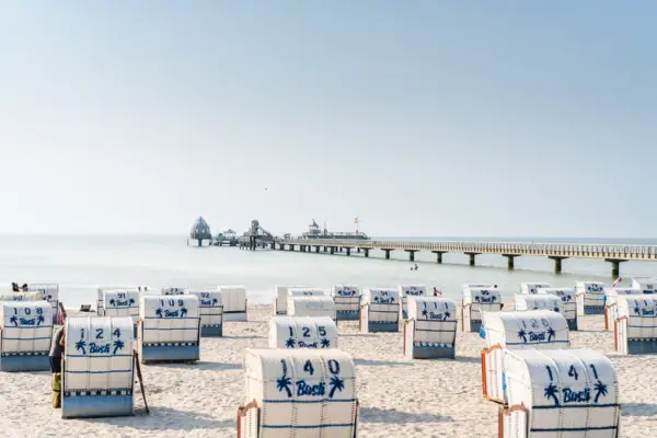 Strand mit weißen Strandkörben und blauen Streifen vor klarem Himmel.