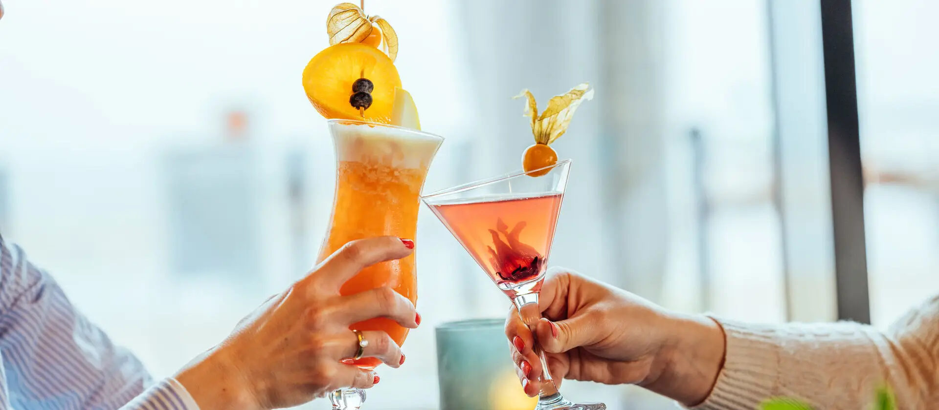 Zwei Menschen stoßen mit zwei orangenen Cocktails mit großer Dekoration am Glasrand an. 