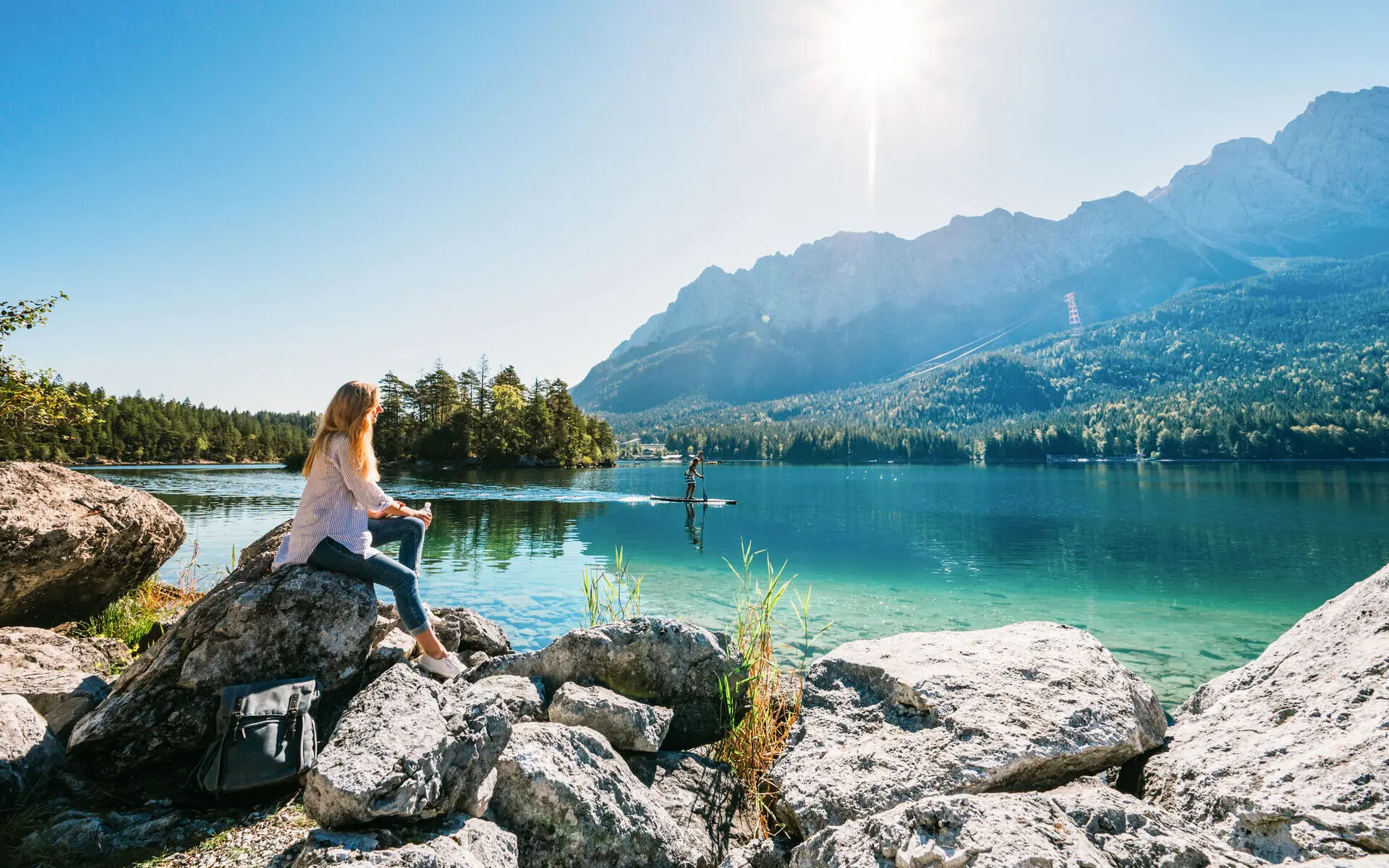 Frau sitzt auf einem Felsen am Ufer eines Sees, umgeben von Bergen und Natur.