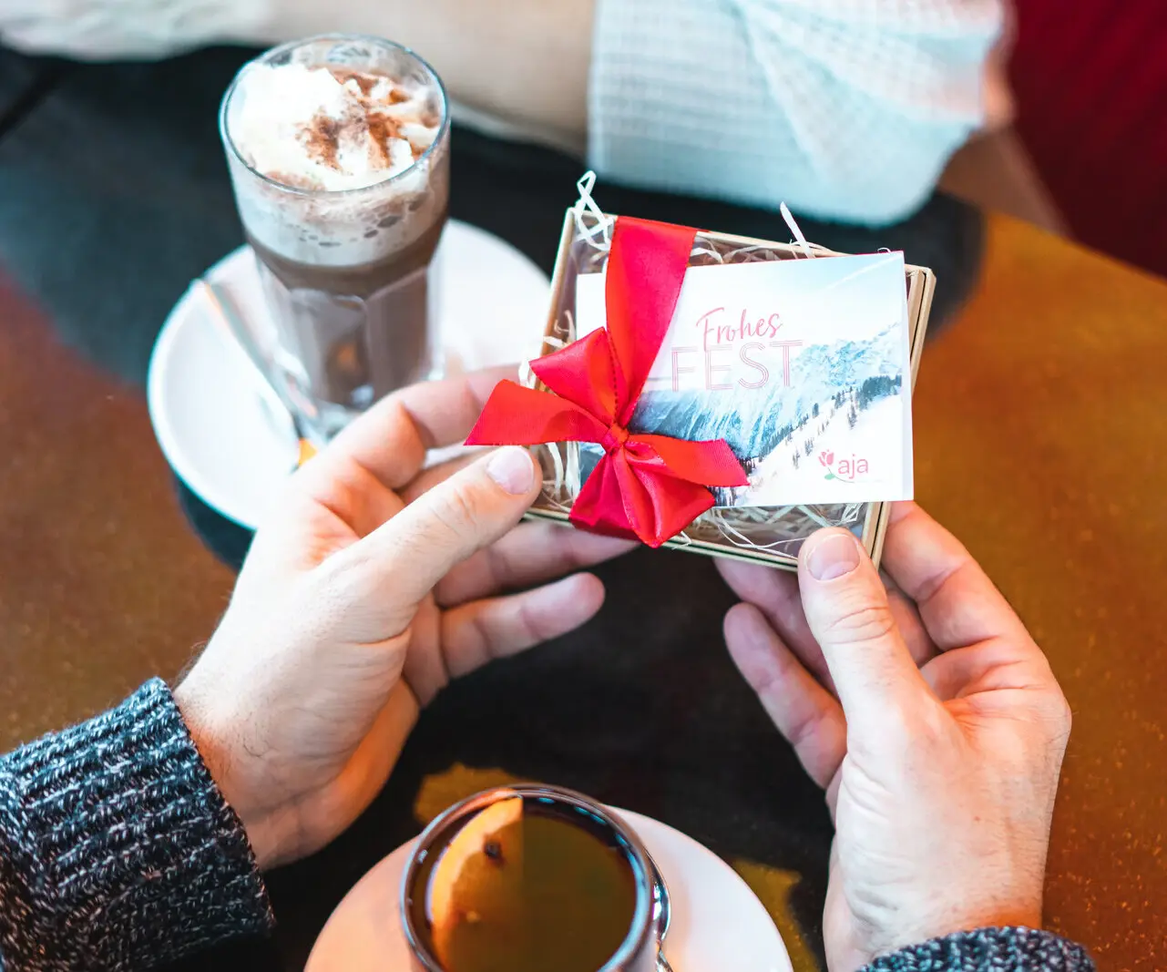 Hände halten eine Geschenkbox mit roter Schleife und eine Tasse Kaffee.