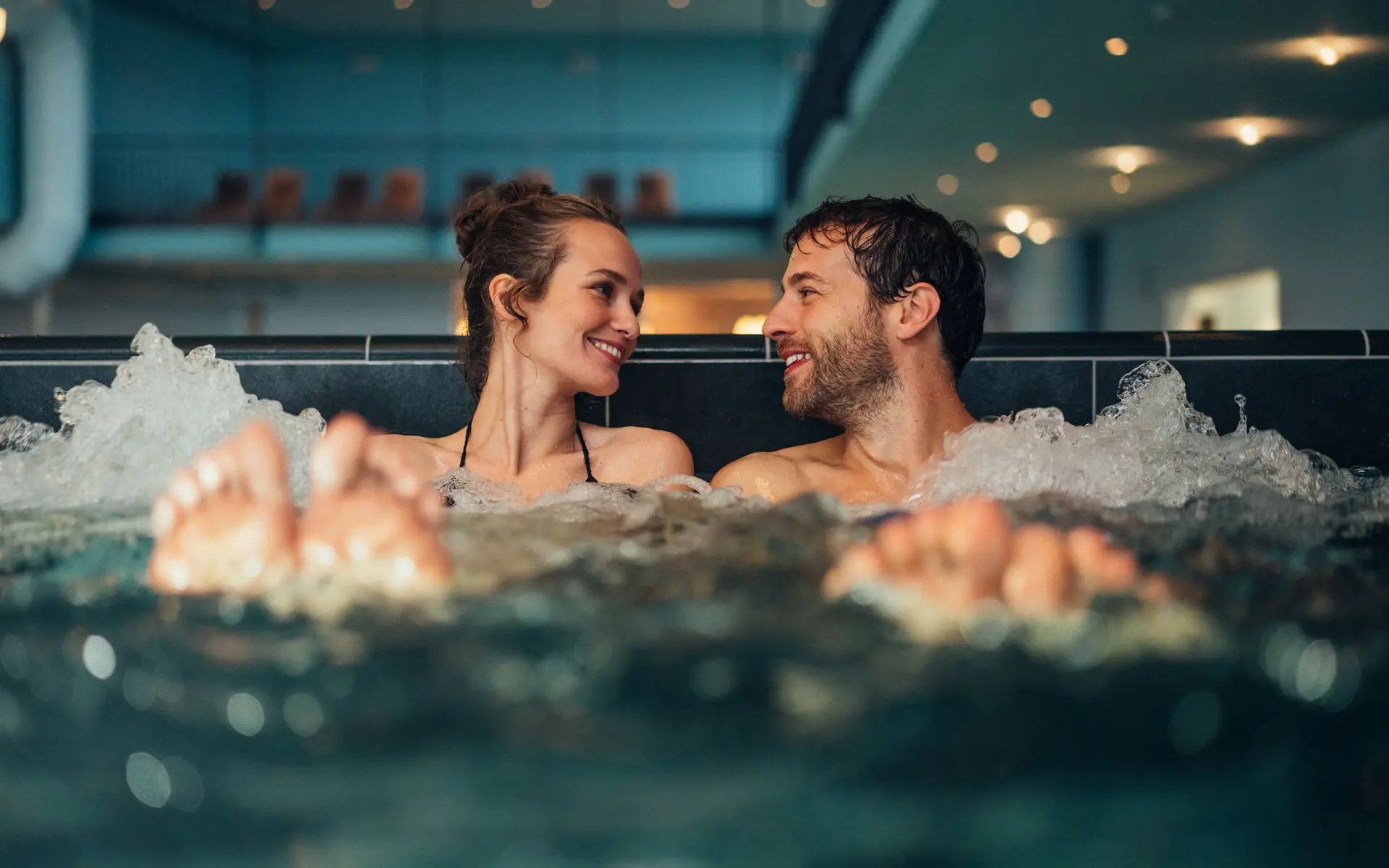 Ein Mann und eine Frau sitzen in einem sprudelnden Schwimmbecken, die Blicke einander zugewandt. Im Hintergrund ist abgedunkelt und leicht verschwommen einige Liegen im oberen Bereich zu erkennen.