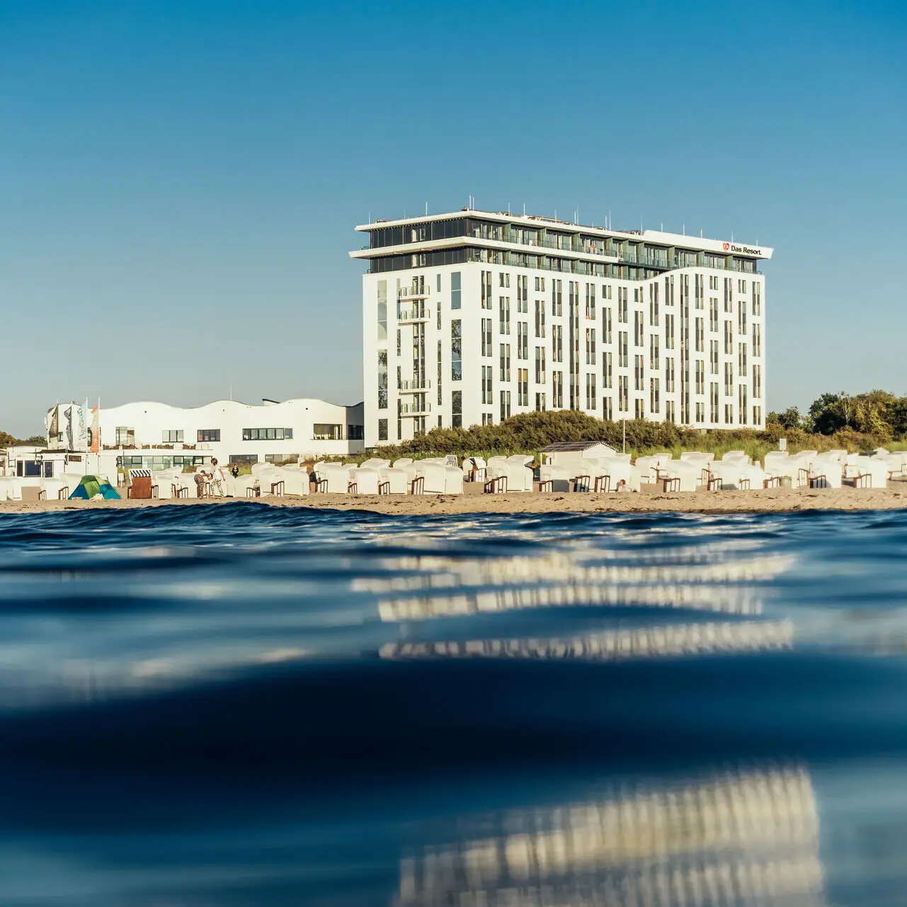 aja Warnemünde Das Hotelgebäude am Strand mit klarem Himmel und Wasser im Vordergrund.