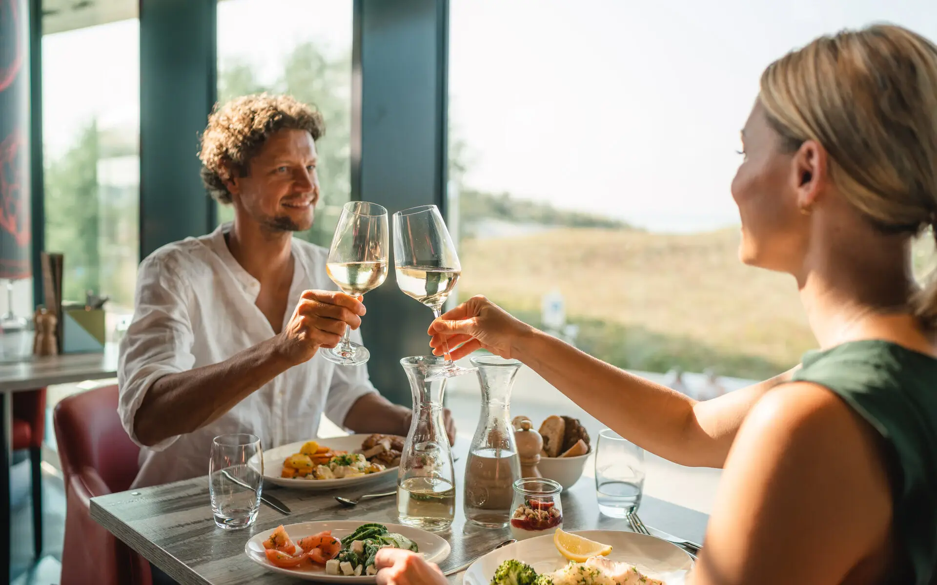 Abendessen im aja Warnemünde Ein Mann und eine Frau stoßen mit Weingläsern an einem Tisch an.