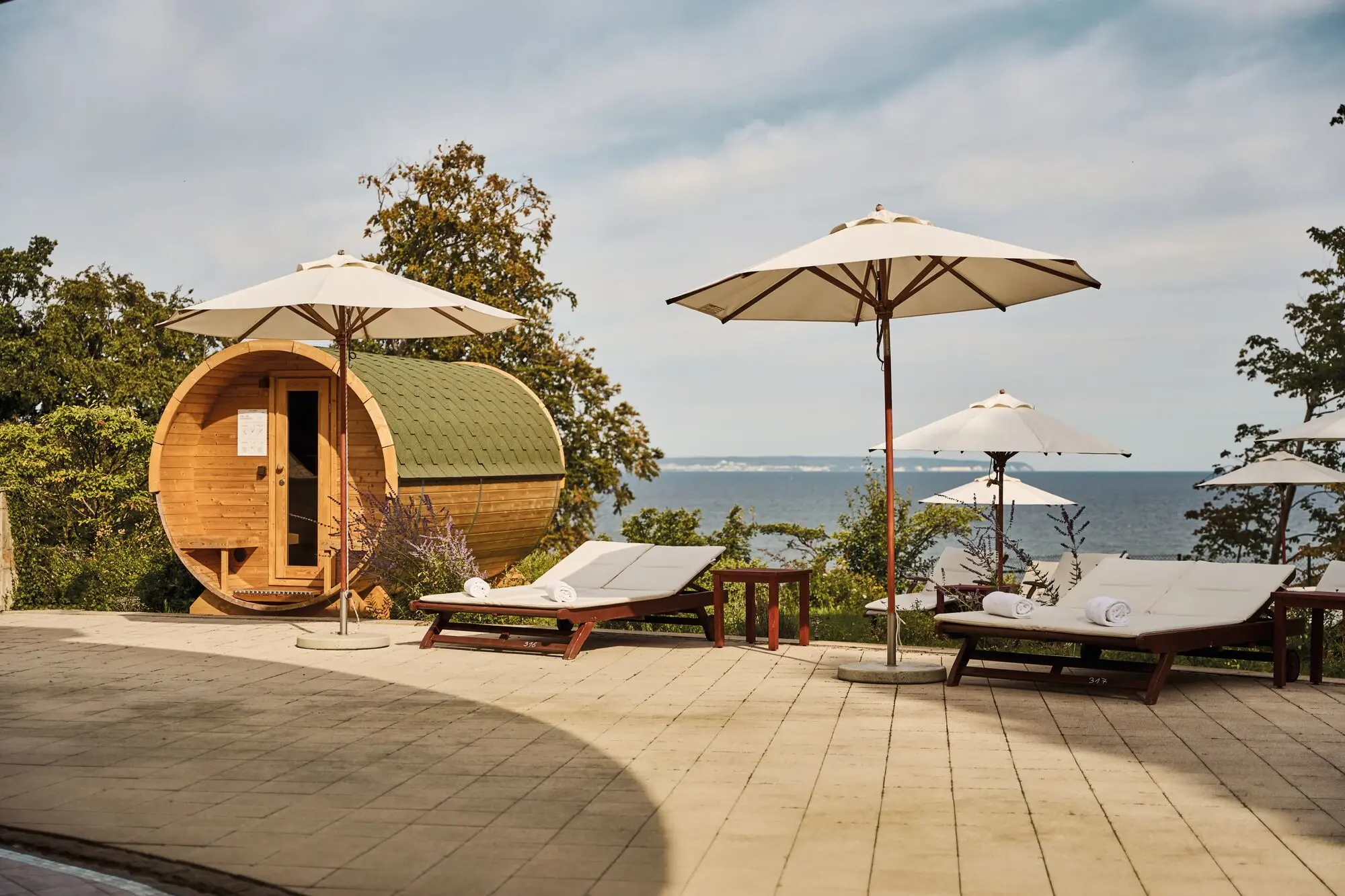 Wellnessbereich mit Außensauna in Fassform, Sonnenliegen und Sonnenschirmen auf einer Terrasse mit Blick auf die Ostsee; ruhige, entspannte Atmosphäre unter blauem Himmel.