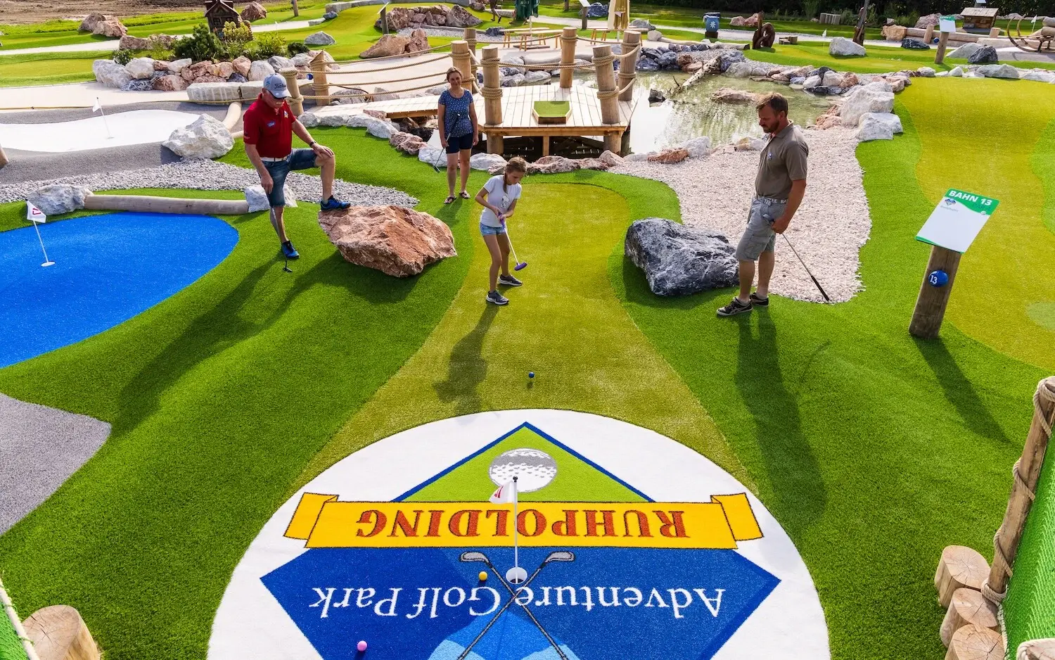 Familie beim Adventure Minigolf im Ruhpolding Adventure Park, umgeben von grünen Kunstrasenbahnen, Felsen und Brücken. 