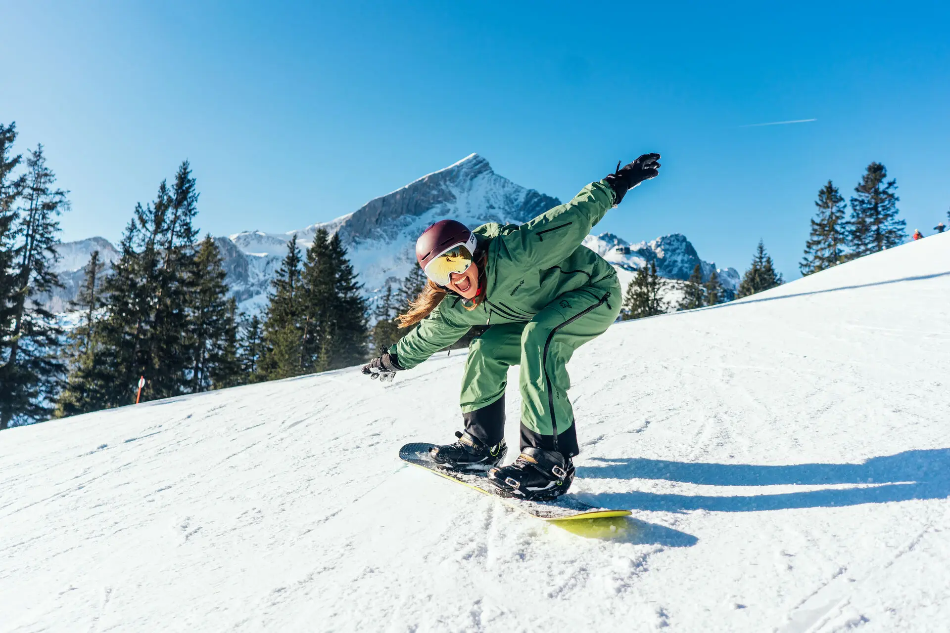 Eine Frau auf einem Snowboard auf einer schneebedeckten Piste streckt die Arme aus und lacht.