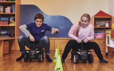 A boy and a girl sit on toy cars and race against each other.