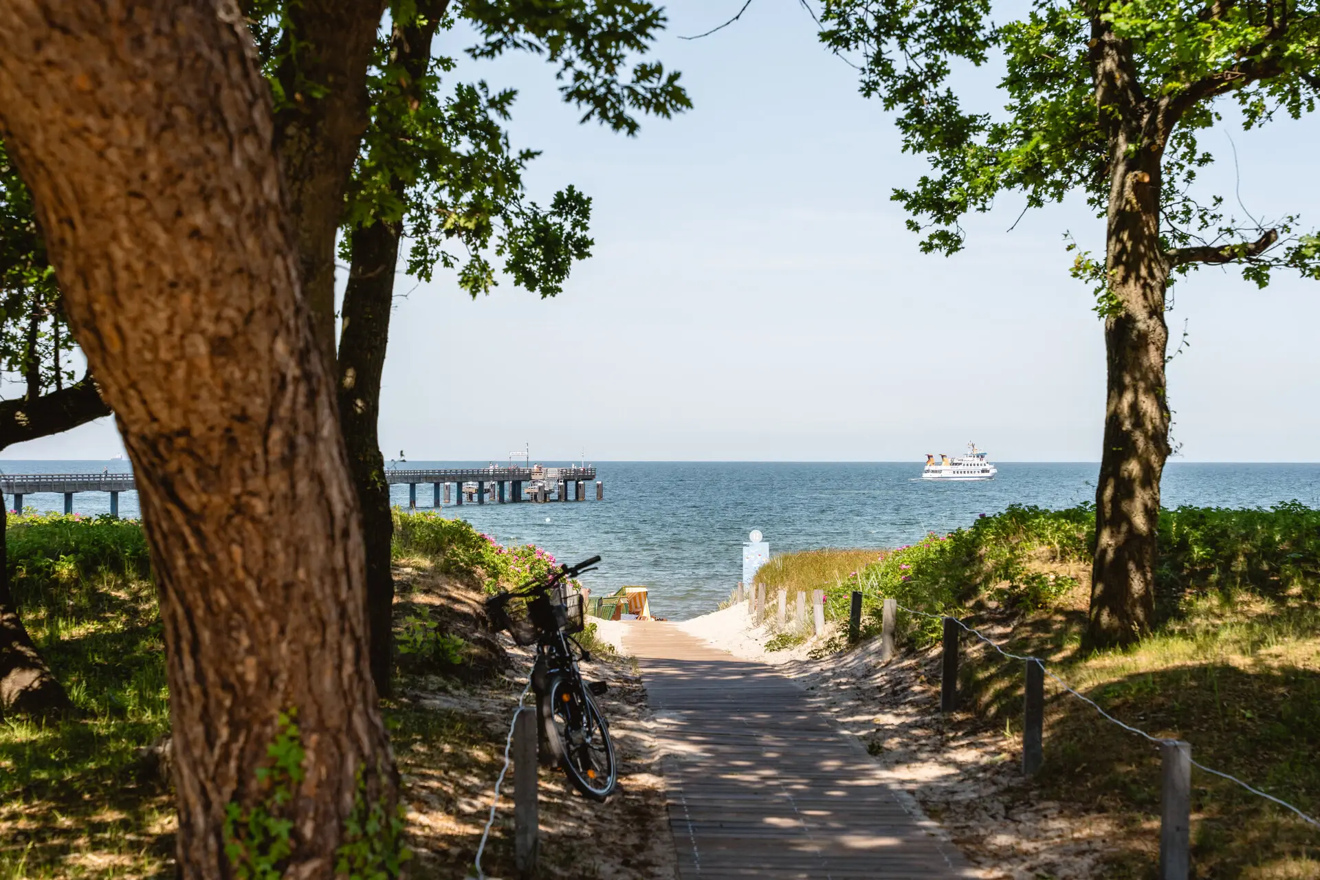 aja Nordperd & Villen Göhren Ein Strand mit Bäumen und Zugang zum Strand, mit Blick auf das Meer, die Seebrücke und ein Schiff.