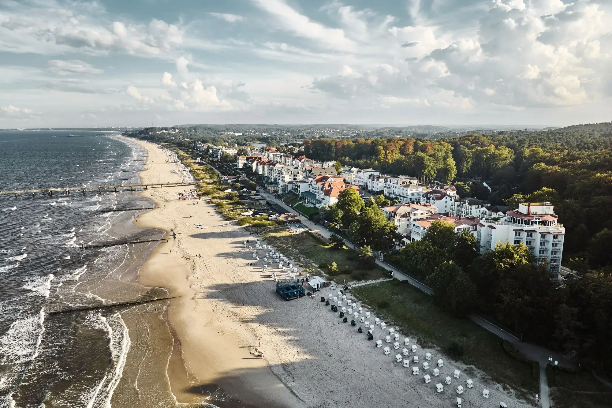 Ein Strand mit Gebäuden und Bäumen, unter einem bewölkten Himmel, mit Blick auf das Meer und umliegende Landschaft aus der Vogelperspektive.