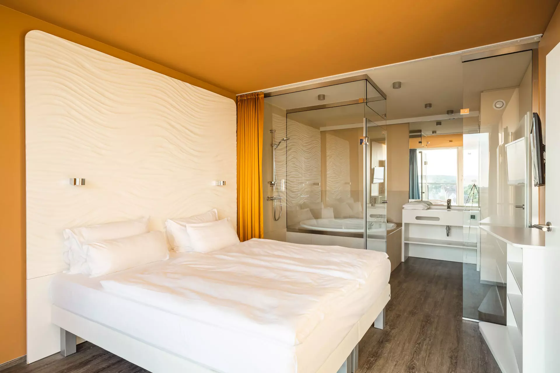 Ein mit weißer Bettwäsche bezogenes Doppelbettsteht vor einer weißen Rückwand mit Struktur. Umrahmt von einer orangenen Wand und einem orangenen Vorhang steht das Bett in einem Raum mit orangefarbener Decke und dunklem Holzboden. Im Hintergrund ist hinter einer gläsernen Trennwand ein Badezimmer zu erkennen.
