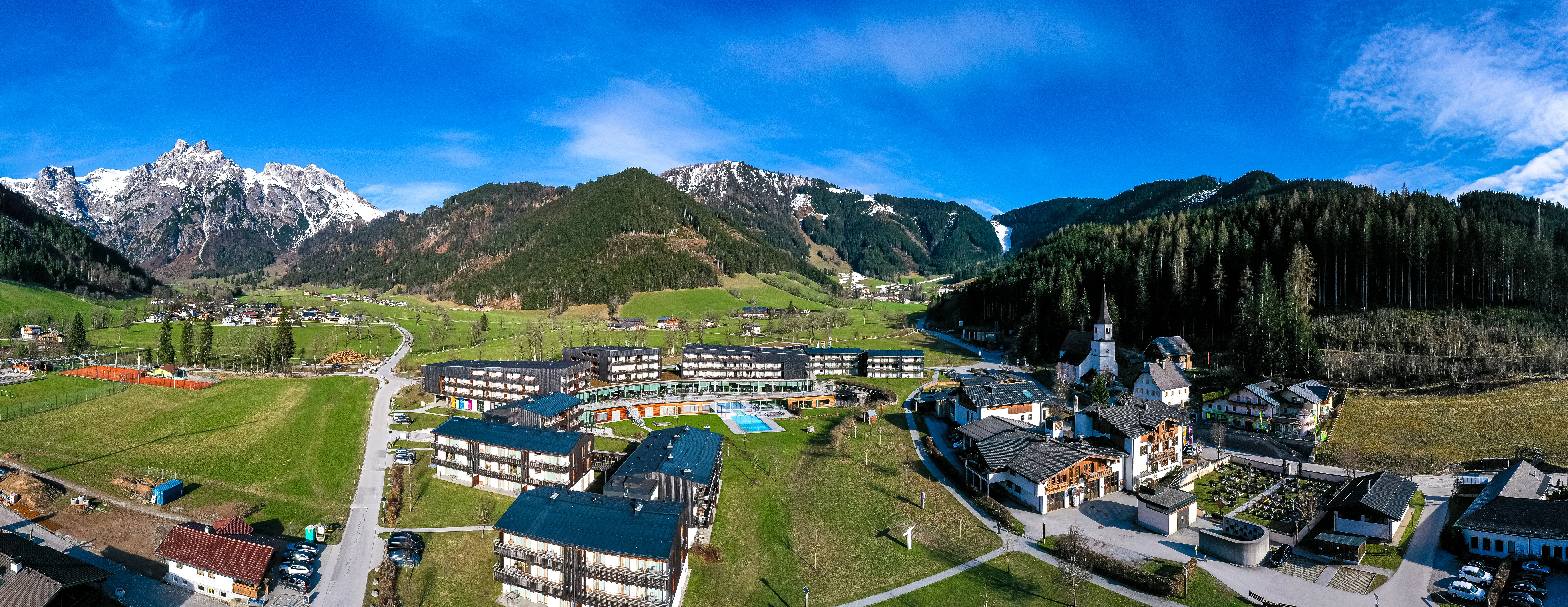 Gruppe von Gebäuden in einem Tal mit Bergen im Hintergrund, umgeben von Bäumen und Gras.