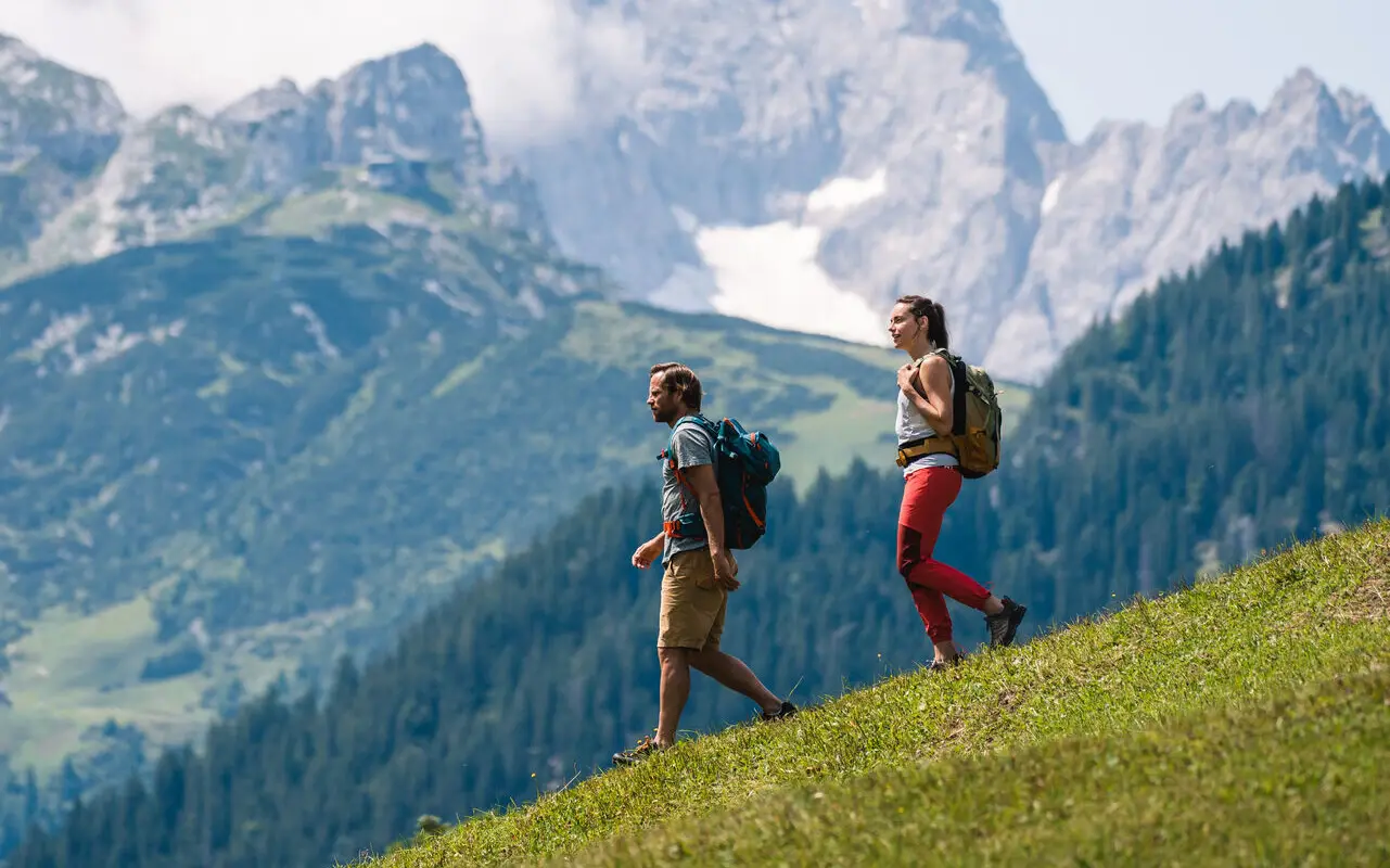 Zwei Personen wandern auf einem Hügel mit Bergkulisse im Hintergrund.