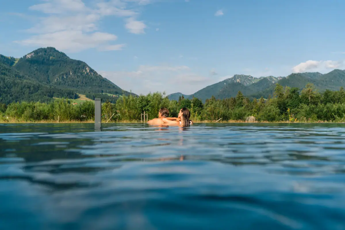 Blick auf die Berge vom Außenpool im aja Ruhpolding Ein Paar schwimmt in einem Pool vor einer Bergkulisse.